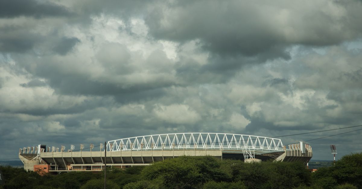 A large modern stadium under a moody cloudy sky with greenery in the foreground.